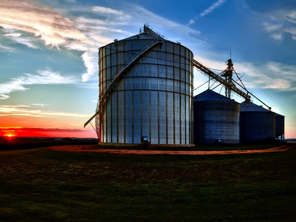 Large grain silos under a dramatic sunset sky, representing agriculture and industry in a rural setting.